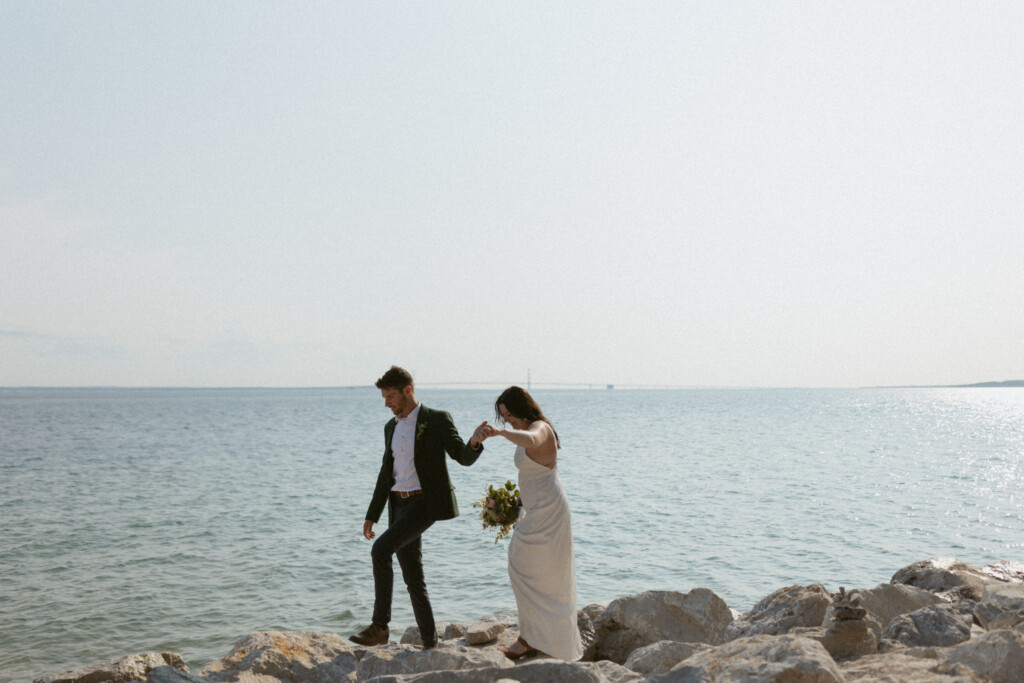 Bride and groom walking along the rocky shoreline of Mackinac Island with the Mackinac bridge in the background.